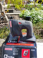 Side view of black and red Craftsman cordless corner sander sitting on its box outdoors. Box shows model 22565 and C3 Cordless branding.