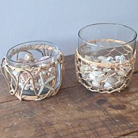 Two glass pots with woven natural fiber netting around them and filled with seashells sitting on a wooden surface.