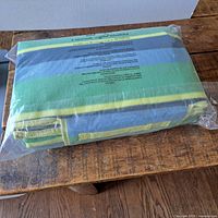 Folded picnic blanket in sealed plastic packaging showing striped green, blue, and yellow pattern on fabric surface lying on wooden surface.