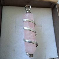 Close-up photo of the rose quartz pendant lying flat on a cushioned surface inside a box, showing the crystal and metal spiral wrap clearly.