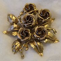 Front view of gold toned roses brooch showing five rose blossoms with leaves and buds on a light background.
