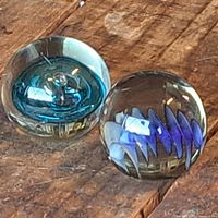 Photo of two round glass paperweights on a wood surface. One is light blue with internal swirls, the other is clear with blue and white zigzag design inside.