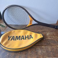 Photo showing the full tennis racket laid on a wooden table next to the yellow zippered cover with Yamaha branding.