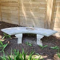 Front angle of curved concrete garden bench on soil with decorative carved edges and two pedestal legs.
