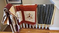 Three blankets displayed on white wooden banister: one patchwork blanket, one quilt with star patterns, and one knitted blanket with yellow border