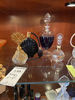 Four glass perfume bottles, including one with gold-tone filigree decoration and black atomizer with bulb spray, displayed on wooden shelf.