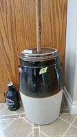 Full view of large two-tone stoneware butter churn crock pot with wooden churn stick and black stoneware bottle on floor.