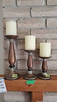 Three wooden and metal candle holders with off-white pillar candles, arranged on a wooden shelf against a brick wall.
