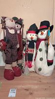 Wide view of four Christmas snowmen standing on wooden floor against an off-white wall. Two tall plush snowmen on left and a lighted snowman and smaller plush snowman on right.