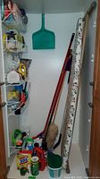 Cleaning supplies including various bottles, green dustpan, mops, brooms, and ironing board with floral cover visible in a closet.
