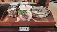 Photo showing wooden tray containing 3 porcelain teacups with intricate floral and geometric design, 3 matching saucers, 3 Denby plates with muted floral design, and a ceramic grape-themed teapot.