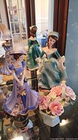 Three porcelain lady figurines, one in lavender gown with feathered hat and bell function, another seated in teal-green dress holding flowers, third in green dress and bonnet. Ceramic floral bouquets with pink roses also visible.