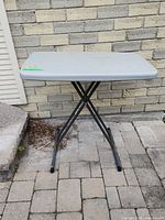 Front angle view of plastic folding table with metal legs against a brick wall on a paved ground