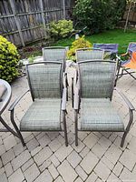 Four garden chairs with metal frames and mesh seats and backs on outdoor patio surface with greenery.