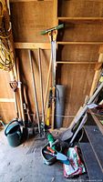 Photo showing several garden tools including rakes, shovels, watering can, and sledge hammer leaning against a wooden wall in a shed.