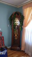 Full view of the wood grandfather clock showing its height and design with decorative greenery around the top.
