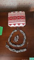 Top view of square ceramic trinket box with red and white Ukrainian folk pattern, showing lid design. Below are a beaded necklace, earrings, and bracelet in black, white, and silver colors arranged on a wooden surface.