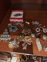 Wide angle view of 15 assorted costume jewellery brooches laid on wooden surface, featuring various metal tones, rhinestones, floral and abstract shapes.