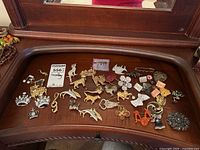Overview of collection of costume jewelry brooches and pins laid out on wooden surface including animal, floral, musical, and crown designs in gold and silver tone finishes.