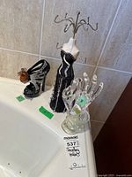 Set of three jewellery holders including a dress form, high heel, and glass hand placed on a white surface against tiled wall.