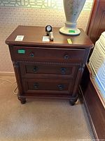 Front view of the wood veneer nightstand showing three drawers with round metal pulls and carved detailing on drawer fronts and legs.