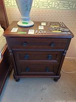 Front view of wood veneer nightstand with three drawers and metal pull handles. A lamp and jewelry are placed on top (not included).