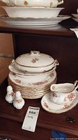 Full view of stacked dinner plates, covered vegetable bowl, salt and pepper shakers, and gravy boat with underplate showing floral rose pattern and gold trim.