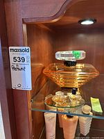 Wide shot showing the large orange plastic perfume display bottle and two small Lancôme Trésor mini perfume bottles on a glass shelf inside a wooden cabinet.
