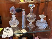 Four glass perfume bottles and one metal perfume atomizer on glass shelf inside wooden cabinet. Bottles vary in height and design.