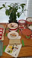 Wide view of the kitchen table displaying a potted plant, decorative plates of multiple patterns and colors, a wooden box, a green ceramic jar with a lid, and kitchen linens.