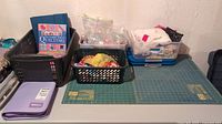 Craft table with Olfa rotary cutting mat, fabric, plastic baskets containing quilting supplies and two quilting books on top of a basket.