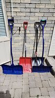 Four snow shovels leaning against a stone wall showing size and color variation.