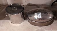 Two cookware items on a concrete floor: a tall stock pot with lid and a roasting pan with domed lid, both stainless steel.