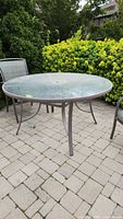 Round outdoor garden table made of plastic top and metal frame, placed on a stone patio with surrounding greenery.