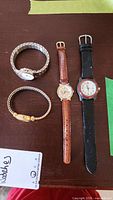 Four wristwatches laid out on a table: a Joanne Rivers gold-tone watch with stones, two Timex watches including a metal stretch band model and a black leather strap model, and a Swiss Army watch with brown leather strap.