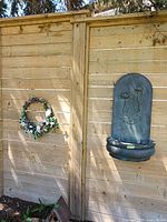 Wide shot showing the floral wreath hanging on wooden fence, the resin water fountain mounted on the fence, the green bird feeder sitting on the ground, and the white floral metal fence decoration leaning against the fence.