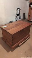 Rectangular wooden blanket box with dovetailed joints and visible wood grain, located in a basement setting.