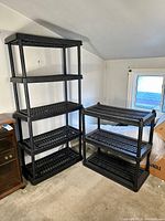 Photo showing two black plastic shelving units: one with five shelves and taller, one with three shelves and shorter, placed against wall near a window.