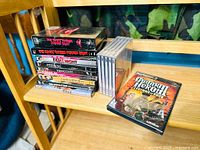 Stack of mostly sealed DVDs and some opened ones on wooden shelving, including two copies of The Rocky Horror Picture Show and a PS2 Guitar Hero III game
