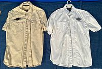 Two shirts hanging side by side: cream on left and white on right, both short sleeve button-up shirts with Harley-Davidson branding.
