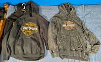Two Harley Davidson hooded sweatshirts laid out flat, showing one black with bright orange logo and one faded black hoodie with similar logo.