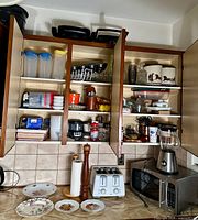 Kitchen cabinets open showing stacked plastic containers with blue/yellow lids, tins with animal silhouettes, various jars and cups, glassware. Countertop shows white toaster, tall wooden pepper grinder, paper towel holder, microwave, and blender.