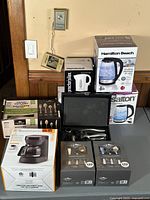 Full lot of kitchen utilities arranged on table including coffee maker, utensil sets, hot water boilers, wine opener set and toaster, all in boxes or cases.