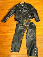 Full view of black leather Harley Davidson motorcycle jacket paired with black leather biker chaps laid out on wooden floor.