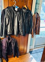 Five jackets hanging on hooks and hangers on a wooden door, showing leather texture and colors black and brown.