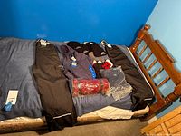 Photo showing two pairs of black snow pants, various winter hats, gloves, and a blue hoodie all laid out on a bed.