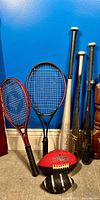 Photo showing two tennis rackets with red and black frames, three baseball bats standing vertically, one silver, one black, one black with a silver handle, and two foam footballs, one red Wilson and one black and white.