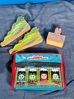 Two wooden mountain track pieces with green and rocky hill designs, one painted wooden castle bridge piece with brick design, plus the blue and red fabric Thomas and Friends carrying case with character images and logo