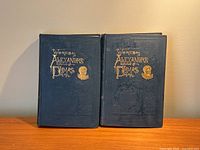 Front view of two antique hardcover books side by side, showing deep blue covers with gilt title 'Works of Alexandre Dumas' and embossed motifs including a portrait.