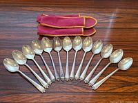 Thirteen spoons arranged in a semicircle on a wood surface with red velvet pouch at top.